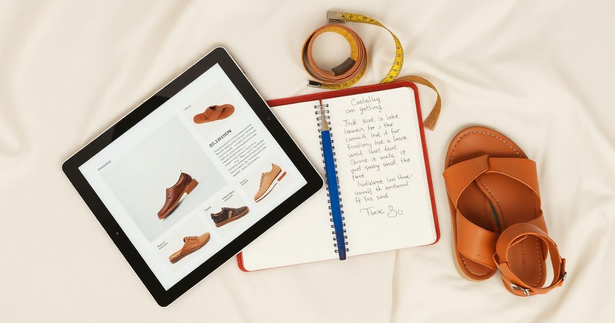 Overhead flat lay of a tablet showing a shoe catalog page next to an open notebook with handwritten notes, a tape measure, and a pair of tan leather sandals on cream linen